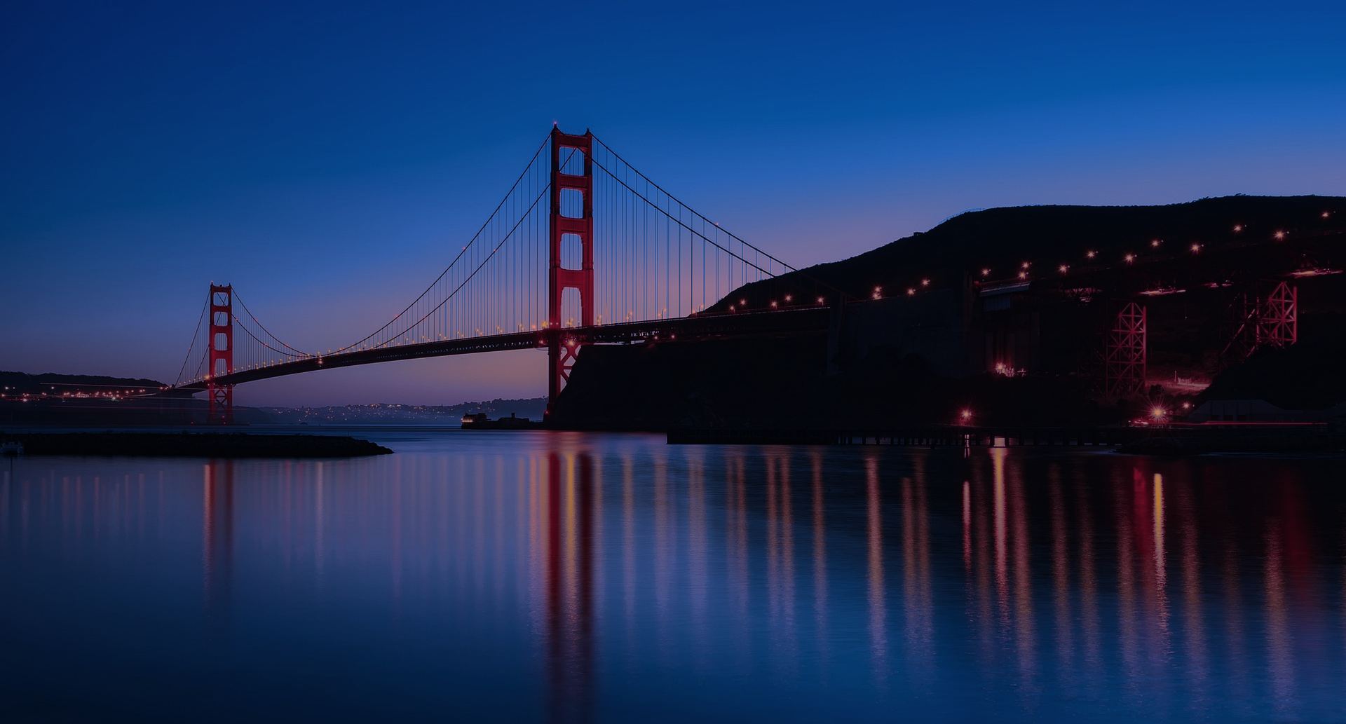 The Golden Gate Bridge at dusk, seen from a San Francisco pier, its lights reflected in the bay.