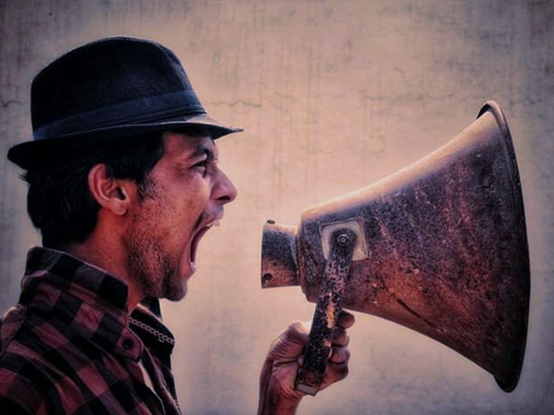 A man in a hat shouting into a big, battered metal megaphone.