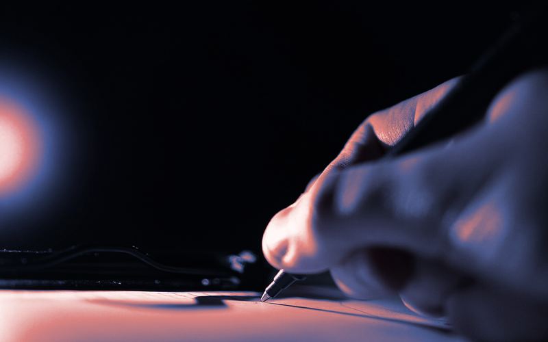 A hand writing with a pen on paper, lit from the side.